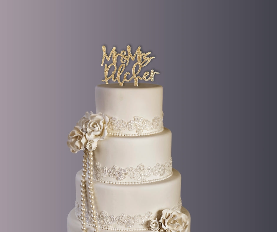 Elegant three-tier white wedding cake with lace details and flowers, featuring glitter gold custom Mr & Mrs name cake topper.