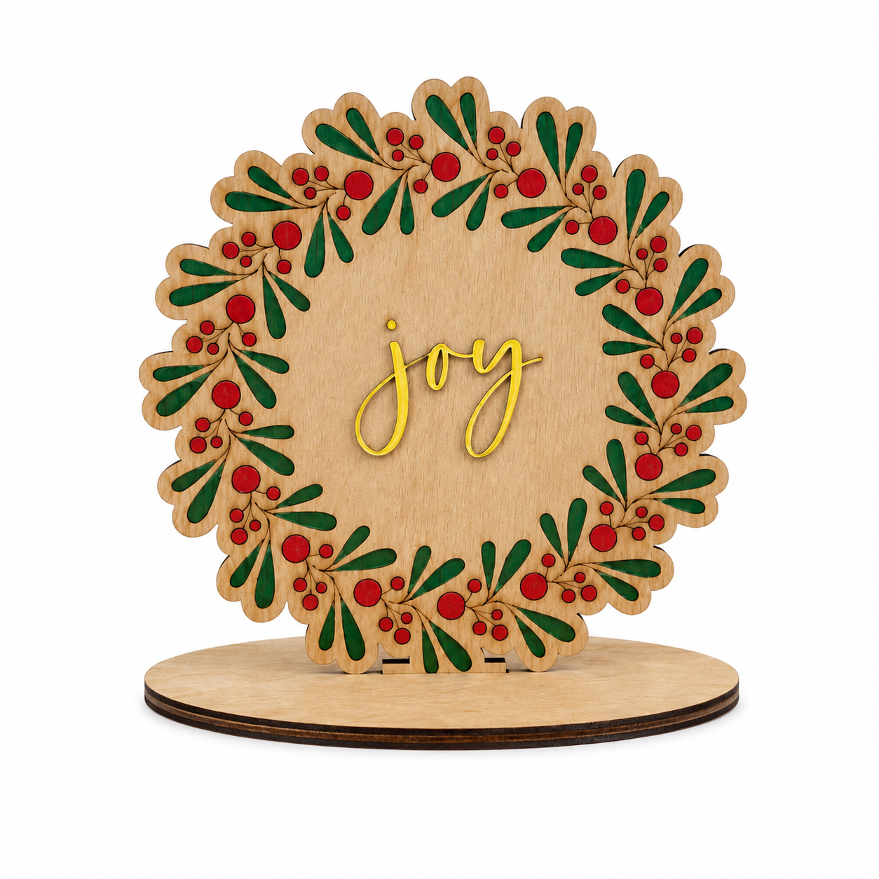 Finished wooden Christmas wreath sign paint kit featuring green foliage, red berries, and a yellow “joy” centerpiece on a tabletop display base.