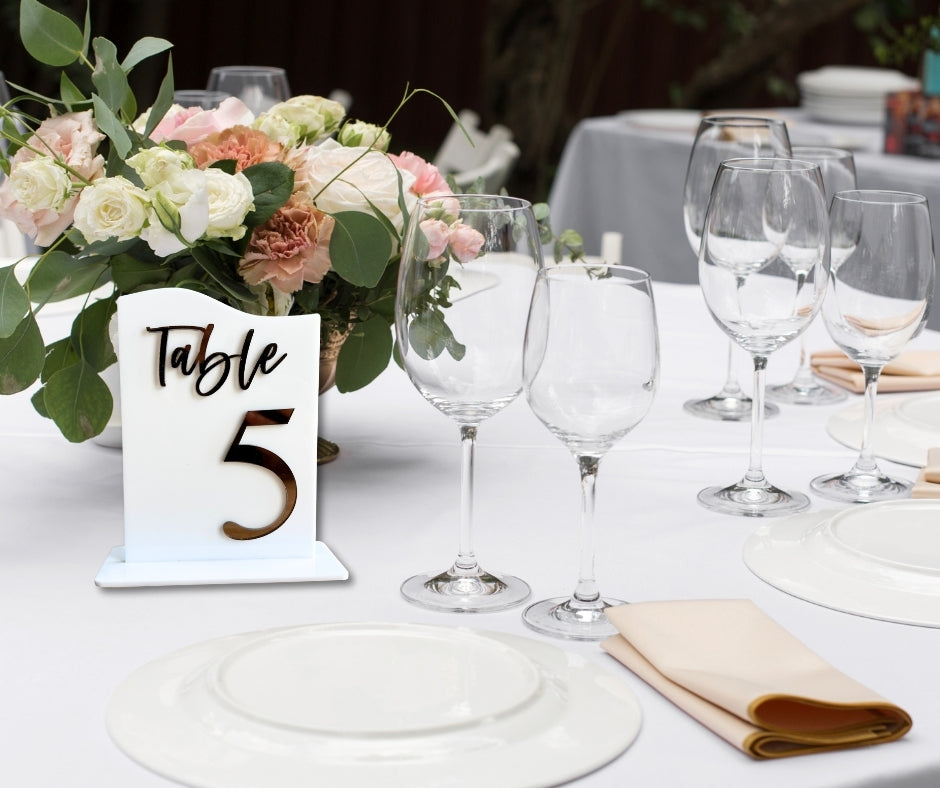 Elegant wedding reception table setting with acrylic table number sign reading Table 5 in gold lettering, surrounded by floral centerpiece and glassware.