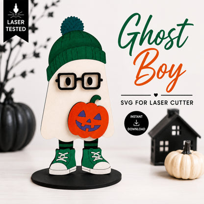 A Halloween-themed wooden cutout of a ghost boy character wearing a green beanie, black glasses, green sneakers, and holding an orange jack-o'-lantern, shown on a white surface with decorative pumpkins and a small house. Text reads 'Ghost Boy', 'SVG FOR LASER CUTTER', and 'Laser Tested'.