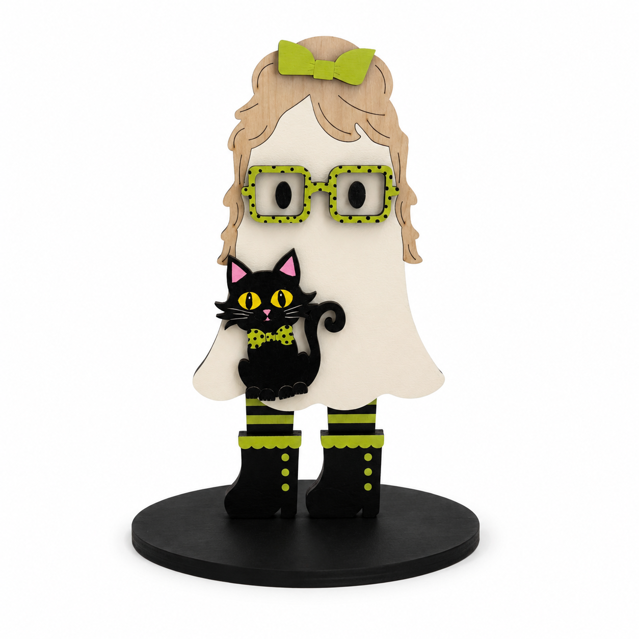 A stylized wooden ghost figurine with blonde hair, green bow, green polka-dot glasses, a black cat on its shoulder, and black boots with green trim, standing on a round black base against a white background.