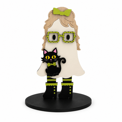 A stylized wooden ghost figurine with blonde hair, green bow, green polka-dot glasses, a black cat on its shoulder, and black boots with green trim, standing on a round black base against a white background.