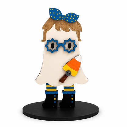 A whimsical wooden ghost figurine with a blue polka-dot bow, blue star-shaped glasses, brown hair, a candy corn accessory, and black boots with blue and yellow stripes, standing on a round black base against a white background.