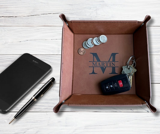 Rawhide leatherette catch-all tray with custom monogram 'M Martin,' holding coins, car keys, and displayed on a white wood background with pen and phone.