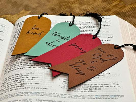 Four leatherette bookmarks with black tassels placed on an open Bible, each engraved with inspirational quotes: “be kind,” “trust & obey,” “pray,” and “give it to God & go to sleep,” in bamboo, teal, red, and brown colors.