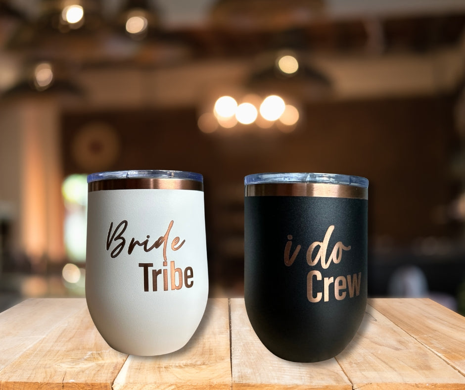 White tumbler engraved with Bride Tribe and black tumbler engraved with I Do Crew, stainless steel wedding party cups with rose gold lettering on wooden table.