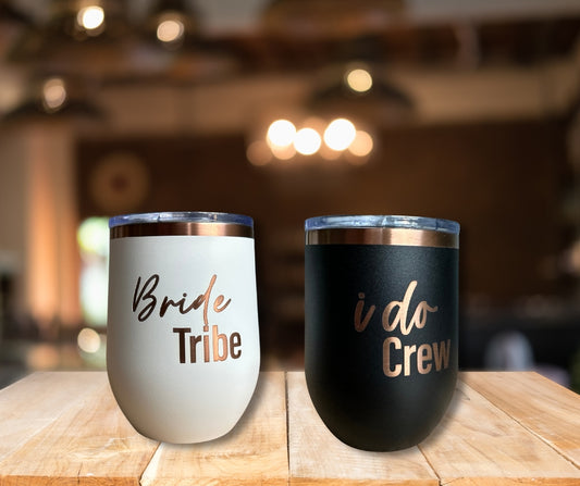 White tumbler engraved with Bride Tribe and black tumbler engraved with I Do Crew, stainless steel wedding party cups with rose gold lettering on wooden table.