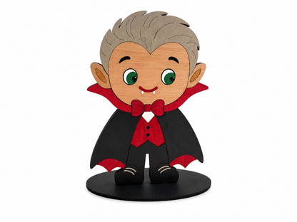 A cartoon-style vampire figurine with gray hair, green eyes, and fangs, wearing a black cape with red lining and a red bow tie, standing on a black circular base against a white background.