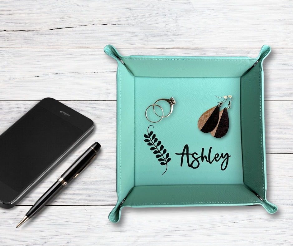 Teal leatherette catch-all tray with personalized name 'Ashley' and leaf design, holding rings and wooden earrings, displayed on a white wood background with pen and phone.