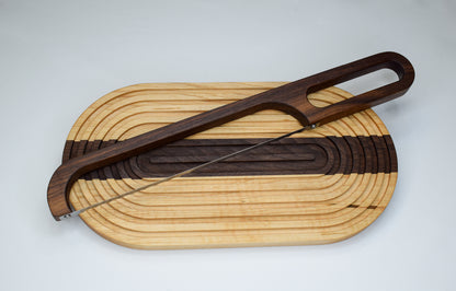 Light colored handmade wooden bread board with a dark colored stripe down the middle and a dark colored bow knife.