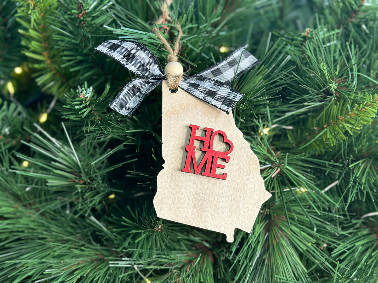 Natural colored wood Christmas ornament in the shape of the state of Georgia and with the word "Home" in red.  Adorned with a black and white buffalo plaid ribbon and wooden bead on twine for easy hanging. 