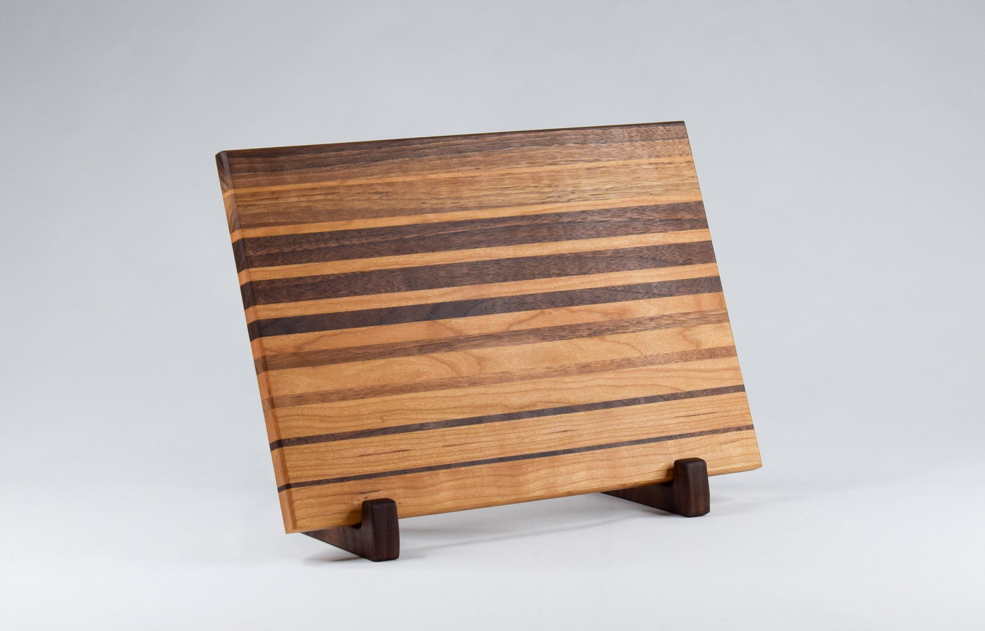 Ombre striped cutting board with alternating walnut and cherry wood. Cutting board displayed on handmade, wooden stand.