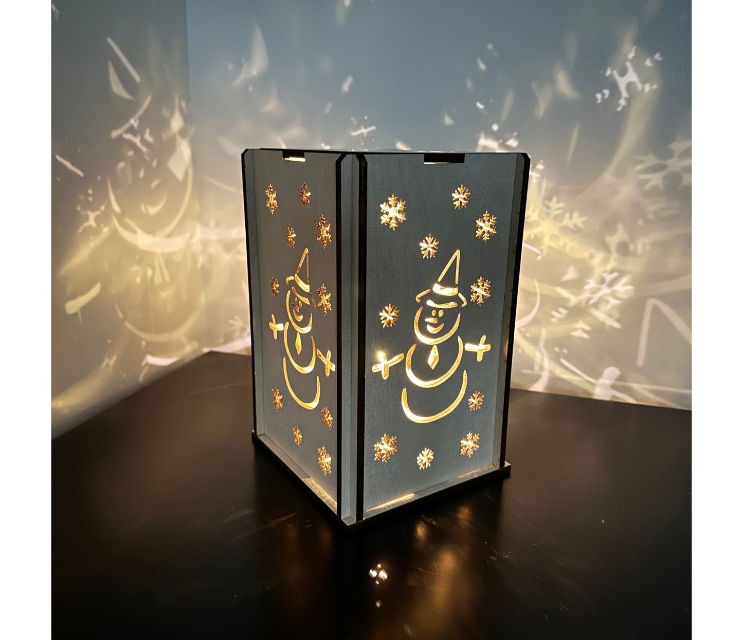 A snowman and snowflake design on a handmade wooden lantern. Lit up and cascading a whimsical light display on the walls.