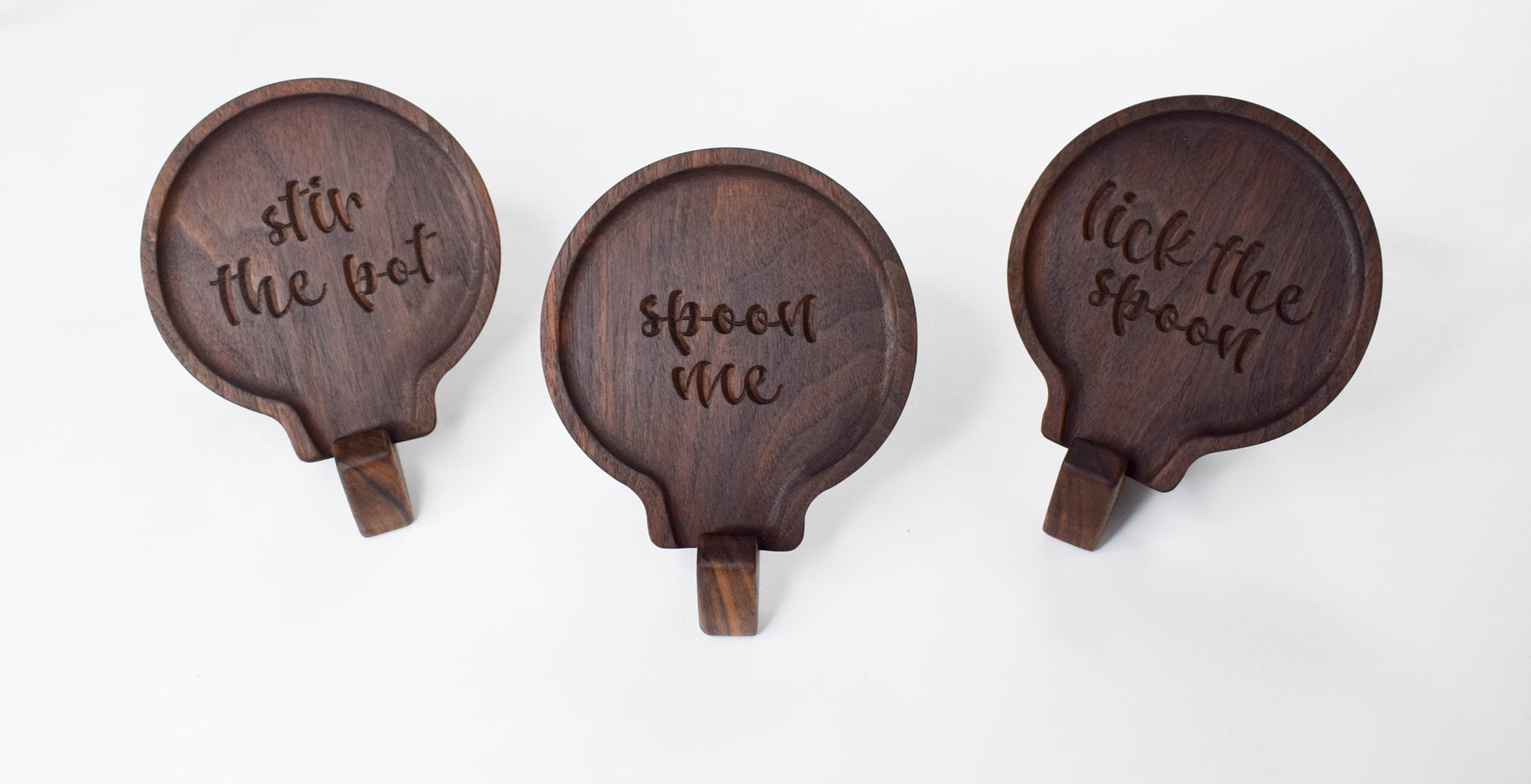 A display of three wooden spoon rests with sayings reading "stir the pot," "spoon me," "lick the spoon." All handcrafted with dark, walnut wood.