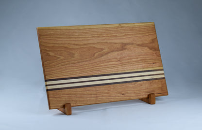 Striped cutting board with alternating stripes of walnut and maple hardwoods against a cherry wood backdrop. Handmade woodworking gift for those who love to cook.