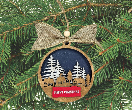 Three layer wooden Christmas Ornament.  Background layer is navy blue with falling snow, middleground shows snow-topped trees, and foreground is a silhouette of a village. Topped with a red banner saying "Merry Christmas." Ornament is finished with twine for hanging, a wooden bead, and a burlap bow.  