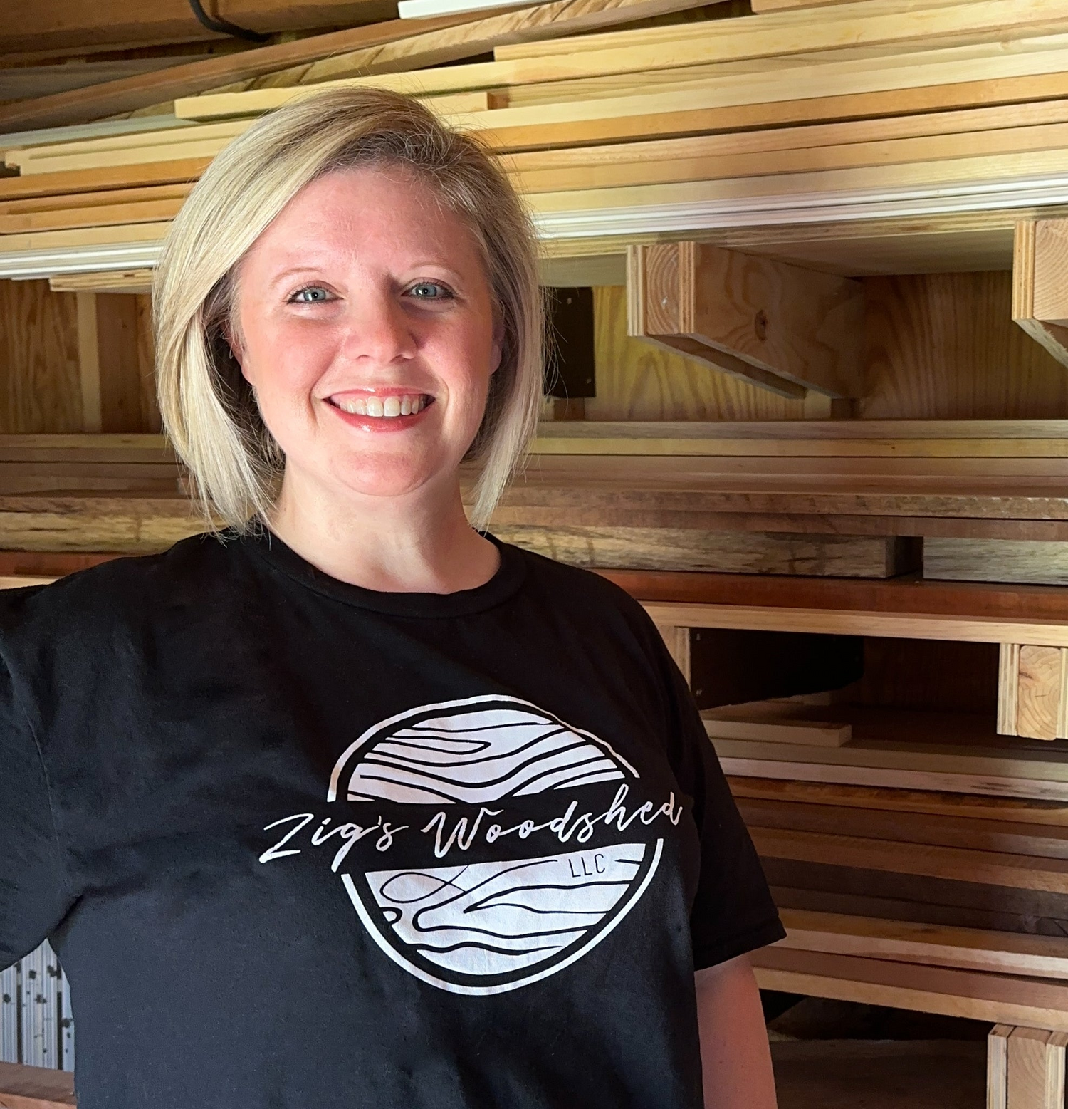 Monica, woodworker and owner of Zig's Woodshed, standing in front of a wall of various types of wood.